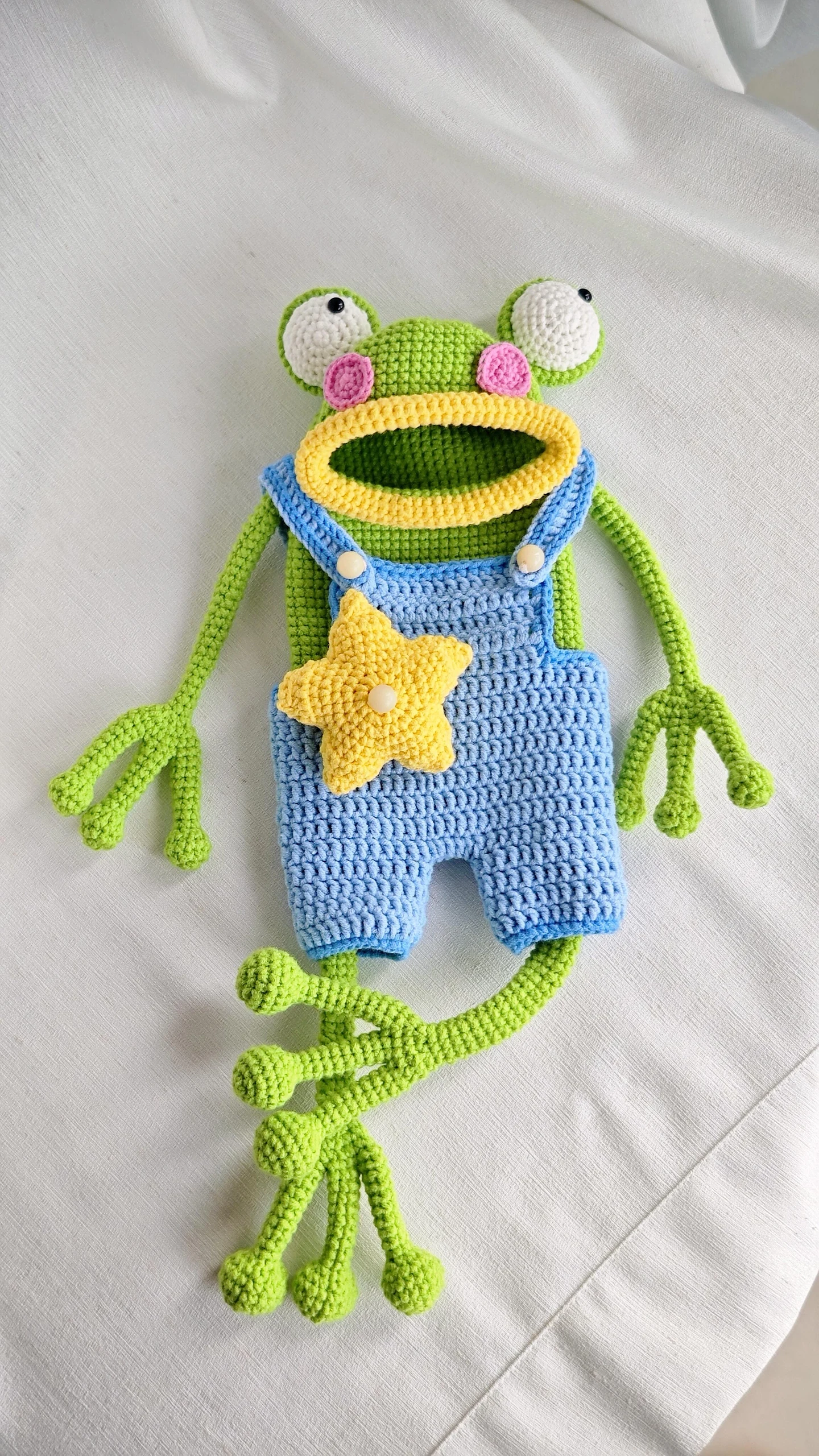 Crochet frog in blue overalls with its long green legs folded together