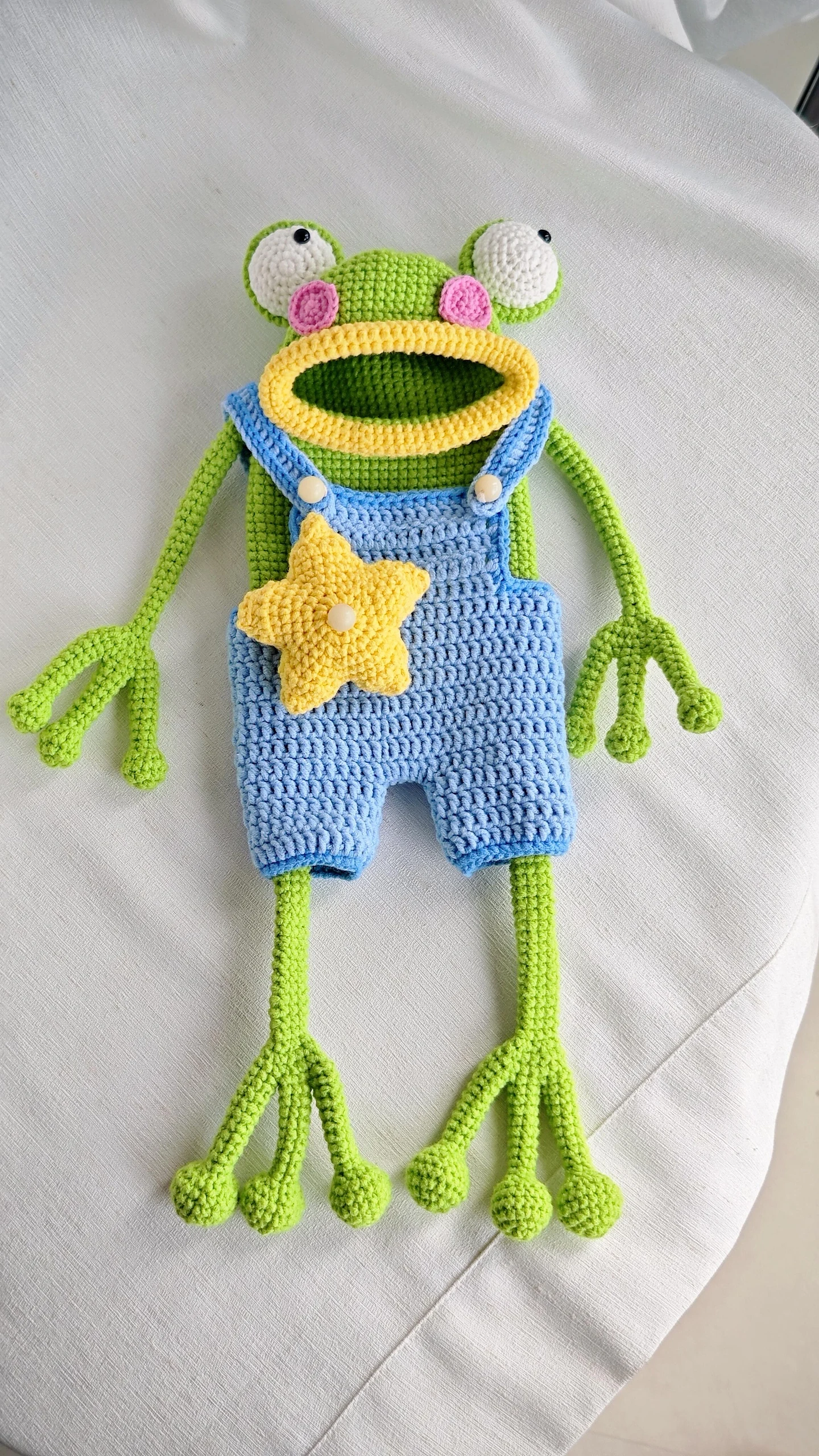 Crochet frog shoulder bag with green limbs, a yellow mouth and star, and blue overalls