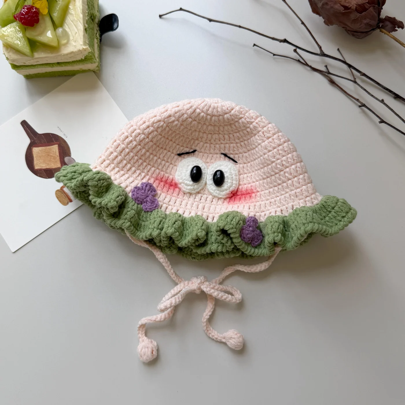 Crochet ruffle hat in pale pink with sage green brim, white eyes and purple flowers